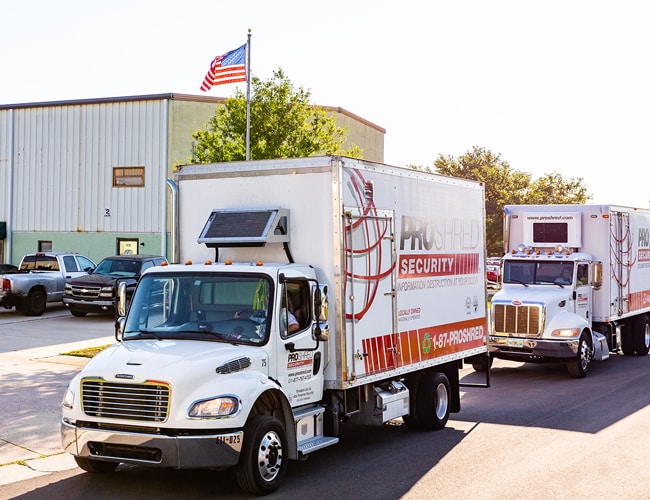 Two PROSHRED mobile paper and document shredding trucks driving beside a building and American flag.